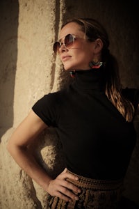 a woman leaning against a stone wall wearing sunglasses