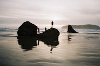 two people standing on rocks on the beach at sunset